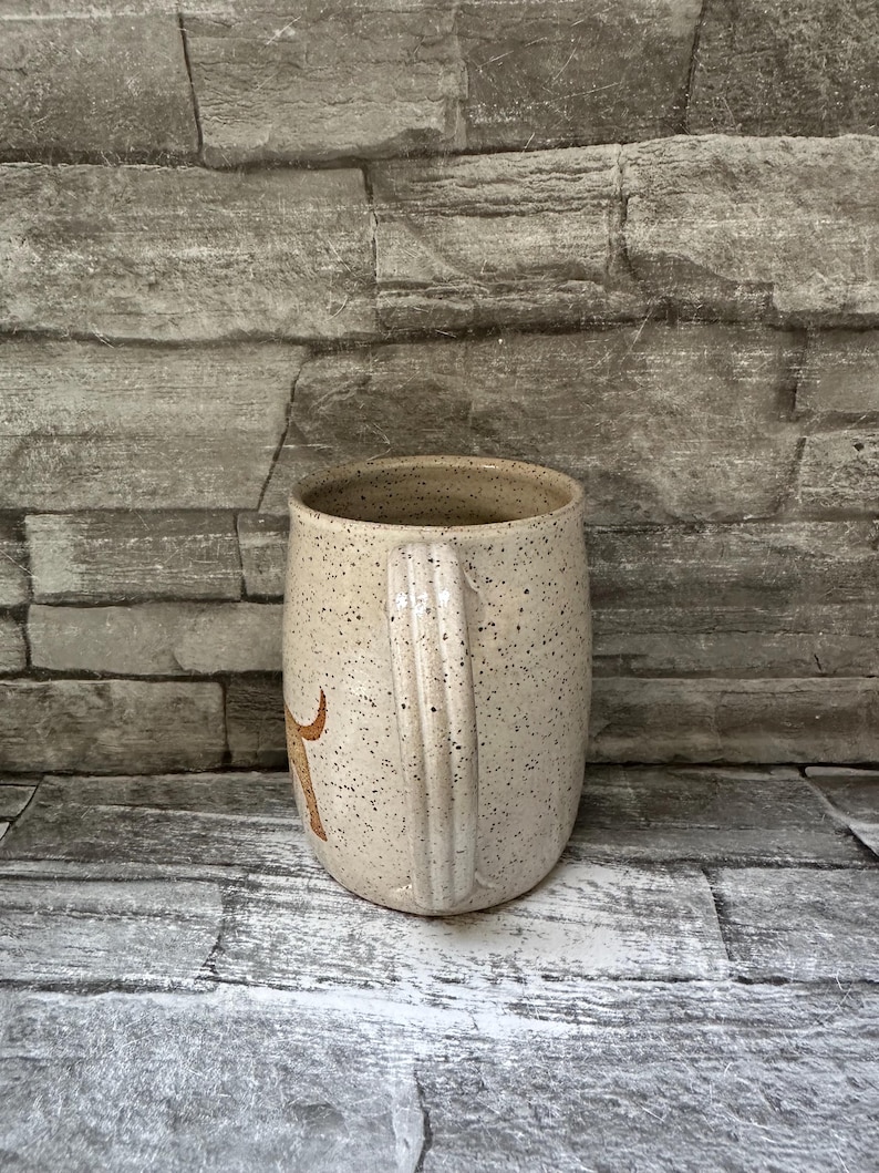 Pode incluir: Uma caneca de cer&acirc;mica bege salpicada com uma al&ccedil;a lisa e arredondada. A superf&iacute;cie externa apresenta uma cor de base creme clara com in&uacute;meras pequenas manchas castanho-escuras distribu&iacute;das por toda a pe&ccedil;a. Um gr&aacute;fico castanho &eacute; vis&iacute;vel.