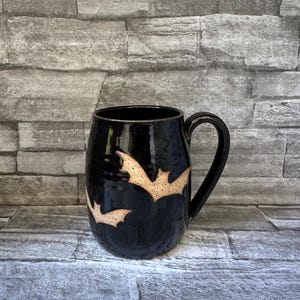 May include: A black ceramic mug with a glossy finish. Two tan-colored bat silhouettes are painted on the side. The mug has a large handle and a rounded shape, perfect for Halloween or spooky season.