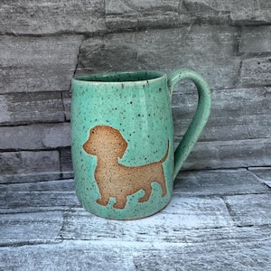 May include: A speckled mint green ceramic mug featuring a brown silhouette of a dachshund dog on the front. The mug has a curved handle and a smooth, glazed finish with dark brown speckles distributed across the entire surface of the vessel.