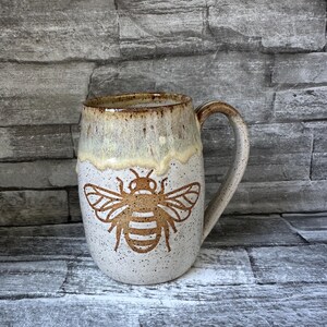 May include: A ceramic mug with a speckled off-white finish and a brown bee illustration on the front. The upper rim features a dripping, cream-colored glaze that transitions into a darker brown edge, complemented by a matching curved handle.