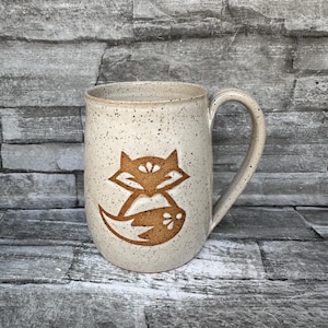 May include: A speckled ceramic mug featuring a brown fox design. The fox is stylized with a geometric shape and a floral accent. The mug has a large handle and a neutral colour palette.