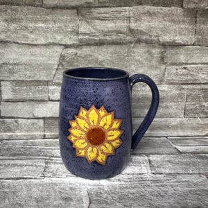 May include: A ceramic mug with a speckled dark blue glaze featuring a hand-painted yellow sunflower design with a brown center. The mug has a rounded body and a curved handle on the right side, showcasing a textured finish throughout the surface.