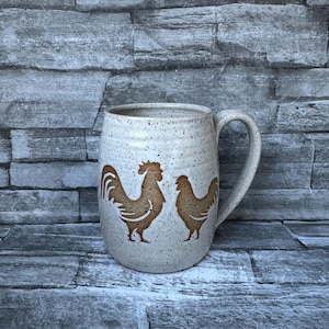 May include: A speckled ceramic mug with a rooster and hen design in brown. The mug has a large handle and a wide opening. The background is a gray stone wall.