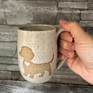 Pode incluir: Uma caneca de cer&acirc;mica com acabamento salpicado em branco sujo, apresentando a silhueta bege de um c&atilde;o dachshund com um pequeno cora&ccedil;&atilde;o recortado no peito. A caneca tem uma asa curva e um corpo arredondado com uma textura r&uacute;stica e salpicada.
