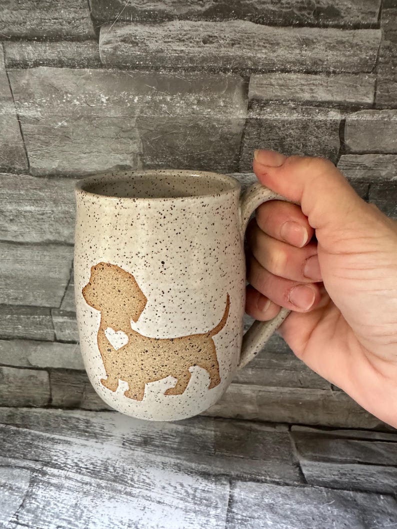 Pode incluir: Uma caneca de cer&acirc;mica com acabamento salpicado em branco sujo, apresentando a silhueta bege de um c&atilde;o dachshund com um pequeno cora&ccedil;&atilde;o recortado no peito. A caneca tem uma asa curva e um corpo arredondado com uma textura r&uacute;stica e salpicada.