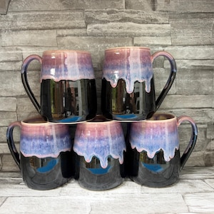 Peut inclure: Cinq tasses en céramique disposées en pile, chacune présentant une base noire brillante et une partie supérieure avec un vernis mélangé violet et rose qui coule vers le centre. Chaque tasse possède une anse incurvée sur le côté.