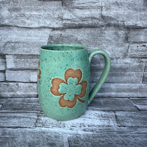 May include: A speckled, turquoise ceramic mug with a brown shamrock design. The mug has a curved handle and a slightly textured surface. The background is a gray stone wall.
