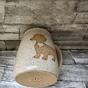Pode incluir: Uma caneca de cer&acirc;mica bege salpicada com a silhueta de um c&atilde;o dachshund com um pequeno cora&ccedil;&atilde;o recortado no peito. A base da caneca exibe um log&oacute;tipo de coroa estampado e o texto "LORA". A caneca tem uma pega curva.
