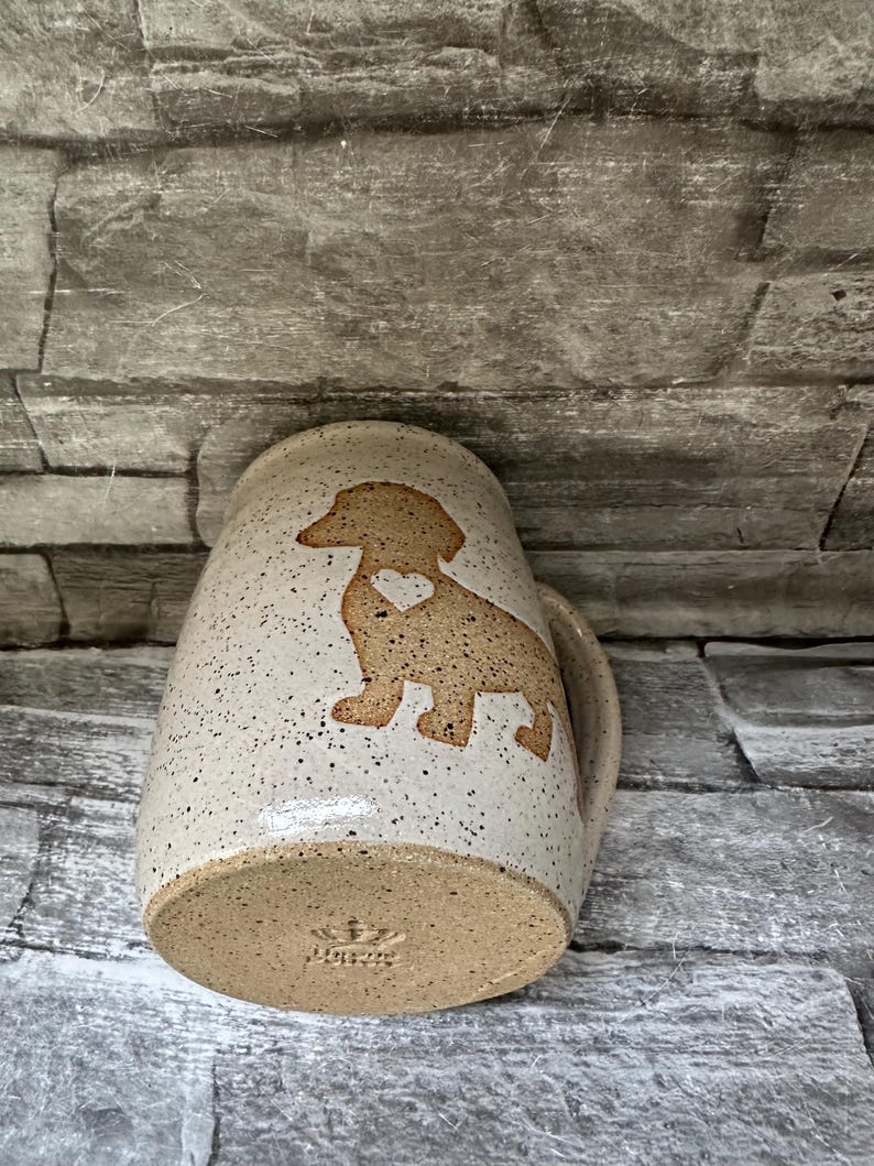 Pode incluir: Uma caneca de cer&acirc;mica bege salpicada com a silhueta de um c&atilde;o dachshund com um pequeno cora&ccedil;&atilde;o recortado no peito. A base da caneca exibe um log&oacute;tipo de coroa estampado e o texto "LORA". A caneca tem uma pega curva.