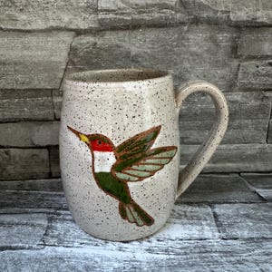 May include: A speckled off-white ceramic mug featuring a hand-painted hummingbird design. The bird has a bright red throat, a white chest, and green wings and tail. The mug has a rounded body and a sturdy handle, with a speckled texture throughout.