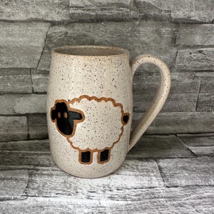 May include: A speckled ceramic mug with a large handle. The mug features a hand-drawn sheep design with a black head and legs, and a white body outlined in brown. The mug is beige with dark brown speckles.