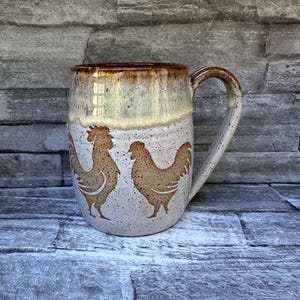 May include: A ceramic mug with a speckled off-white base and a brown glazed rim. The body of the mug features two brown rooster silhouettes facing each other. It has a single curved handle on the right side and a smooth, rounded cylindrical shape.