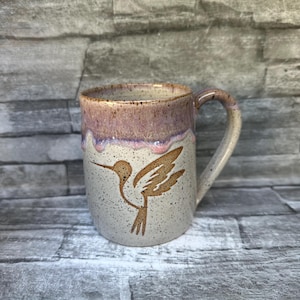 May include: A ceramic mug with a speckled cream-colored body featuring a brown hummingbird silhouette. The upper rim has a drippy, lavender-toned glaze that overlaps the cream base. The mug includes a curved handle and a smooth, rounded opening.