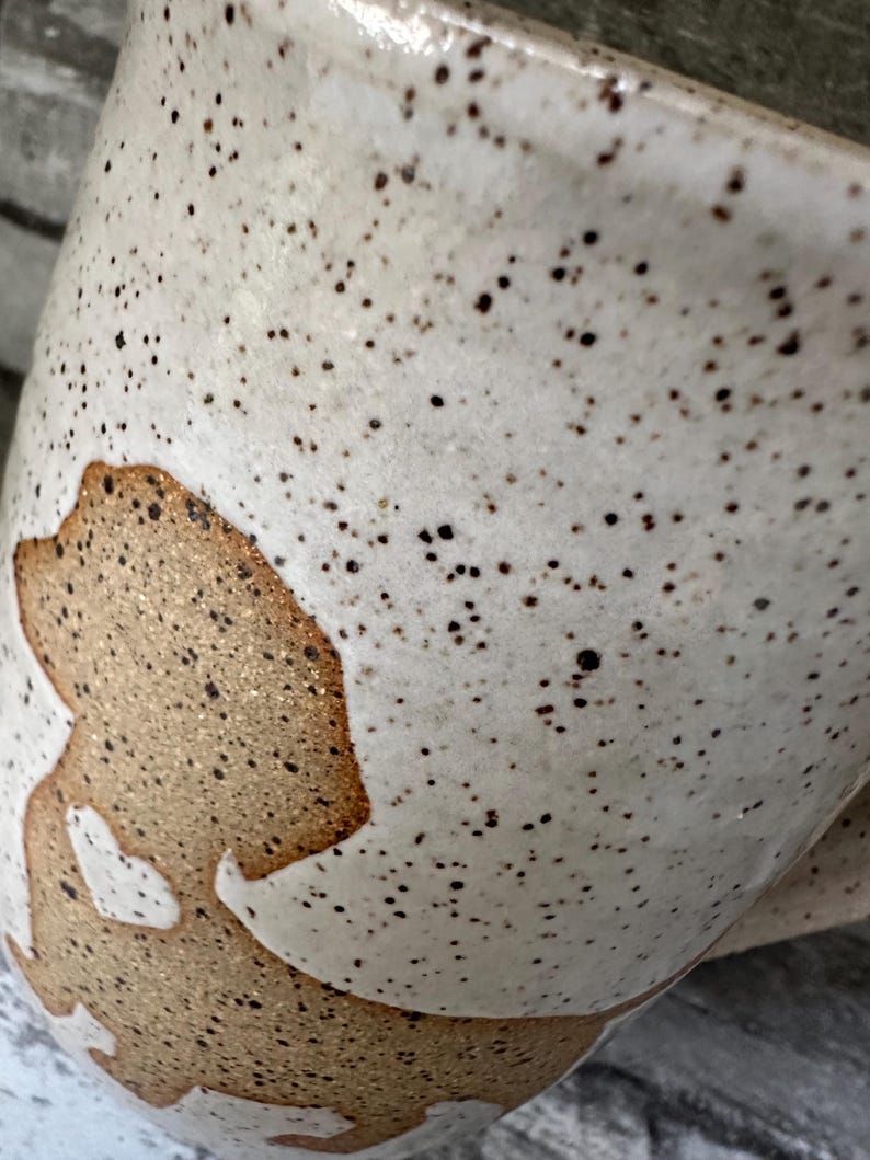 Pode incluir: Uma caneca de cer&acirc;mica com esmalte esbranqui&ccedil;ado salpicado, apresentando uma silhueta de c&atilde;o lisa e sem esmalte na cor castanho-claro na lateral. A silhueta do c&atilde;o cont&eacute;m um pequeno recorte branco em forma de cora&ccedil;&atilde;o. Caneca curva e brilhante.