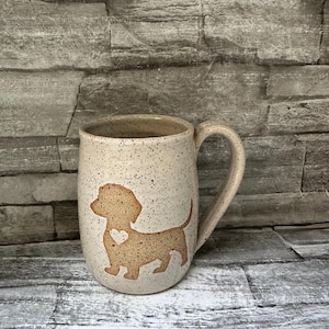 Pode incluir: Uma caneca de cer&acirc;mica cor de creme com pintas, apresentando a silhueta castanha de um c&atilde;o dachshund na parte frontal. O design do c&atilde;o inclui um pequeno recorte em forma de cora&ccedil;&atilde;o no peito. A caneca tem uma asa curva e corpo arredondado.