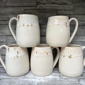 May include: A set of five ceramic mugs with a smooth, cream-colored glaze and a distinctive drip pattern in shades of brown and tan around the upper rim. Each mug features a rounded body, a sturdy side handle, and a glossy finish throughout.