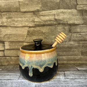 Peut inclure: Un pot à miel en céramique au corps arrondi, doté d'une base noire et d'un motif de glaçure dégoulinante bleu clair et beige. Le pot comprend un couvercle noir assorti avec un bouton central et une cuillère à miel en bois clair.