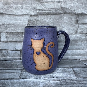 May include: A ceramic mug with a dark blue speckled glaze. The mug features a tan cat silhouette with a heart detail. The handle and rim are also dark blue. The mug is designed for beverages.