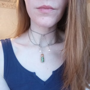 Puede incluir: Un collar gargantilla de cadena plateada con un pequeño colgante de vial de vidrio lleno de purpurina verde y dorada. El collar tiene múltiples hebras y pequeños dijes colgantes transparentes.