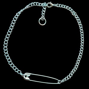 May include: Silver-toned chain necklace with a large safety pin pendant. The necklace features a curb chain design and an adjustable length. The safety pin is positioned at the front of the necklace, creating a unique and edgy look against a black background.