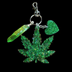 May include: A keychain with a green glitter marijuana leaf, a green glitter heart, and a green crystal. The keychain has a silver clasp and a silver key ring. The background is black.