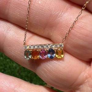May include: A gold necklace featuring a bar pendant with six oval-cut gemstones in a rainbow of colors: light blue, orange, pink, blue, and yellow. The bar is accented with small, clear gemstones. The necklace is held in a hand against a blurred green background.