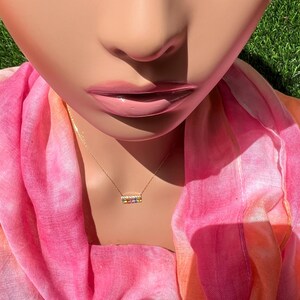 May include: A gold-colored necklace with a rectangular pendant featuring a row of colorful gemstones. The necklace is displayed on a mannequin wearing a pink and orange scarf. The background is a close-up of a face and green grass.