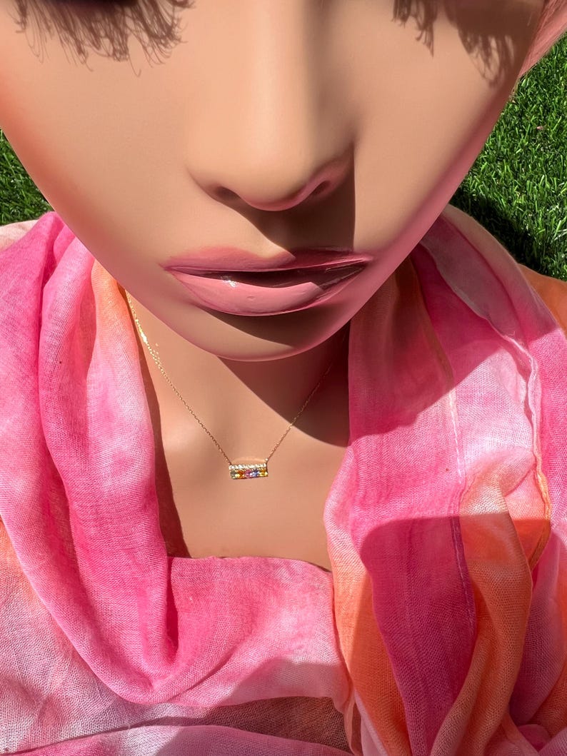 May include: A gold-colored necklace with a rectangular pendant featuring a row of colorful gemstones. The necklace is displayed on a mannequin wearing a pink and orange scarf. The background is a close-up of a face and green grass.
