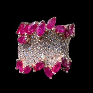 May include: A rose gold ring with a band encrusted with small, clear gemstones. The ring is accented with marquise-cut, vibrant pink gemstones around the edges. The ring is set against a black background.