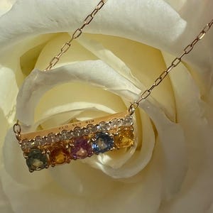 May include: A gold necklace with a bar pendant featuring a row of colorful gemstones and small diamonds. The necklace is set against a soft white background, highlighting the jewelry's details. The pendant has the text "K18 1.15 0.17".