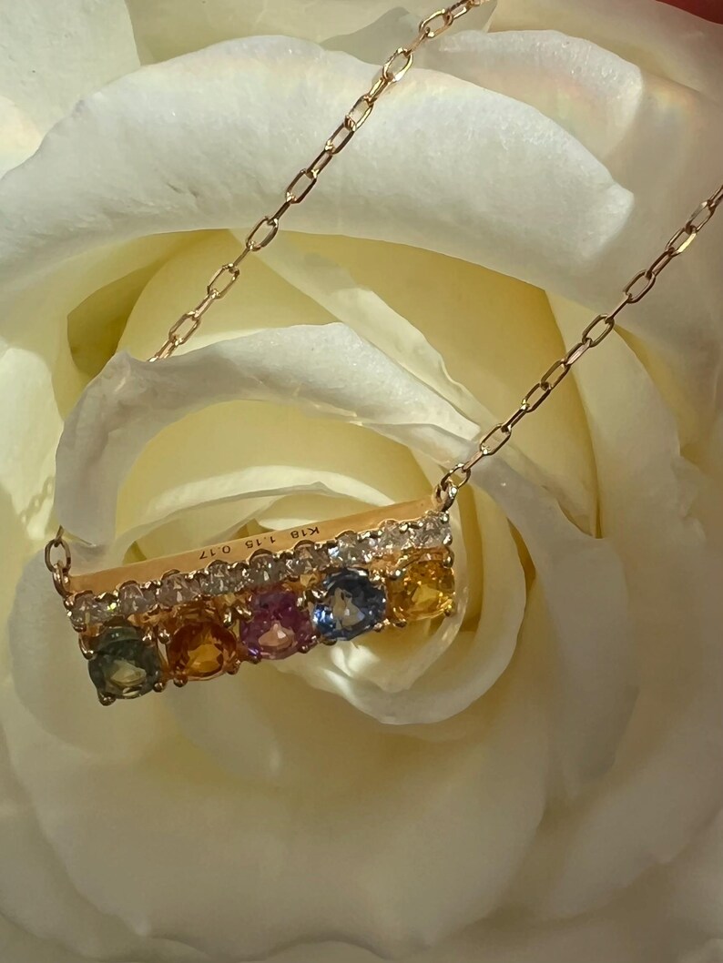 May include: A gold necklace with a bar pendant featuring a row of colorful gemstones and small diamonds. The necklace is set against a soft white background, highlighting the jewelry's details. The pendant has the text "K18 1.15 0.17".