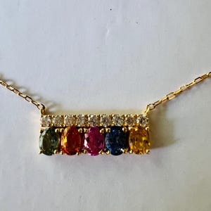 May include: A gold necklace featuring a bar pendant with six oval-cut gemstones in a rainbow of colors: green, orange, pink, blue, and yellow. The bar is accented with a row of small, clear gemstones. The necklace chain is delicate.