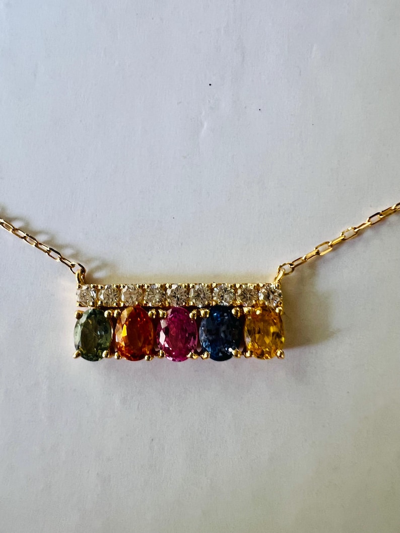 May include: A gold necklace featuring a bar pendant with six oval-cut gemstones in a rainbow of colors: green, orange, pink, blue, and yellow. The bar is accented with a row of small, clear gemstones. The necklace chain is delicate.