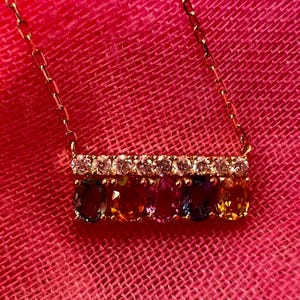 May include: A gold necklace with a bar pendant featuring a row of clear stones above a row of oval-cut gemstones in various colors. The necklace is displayed against a pink background.