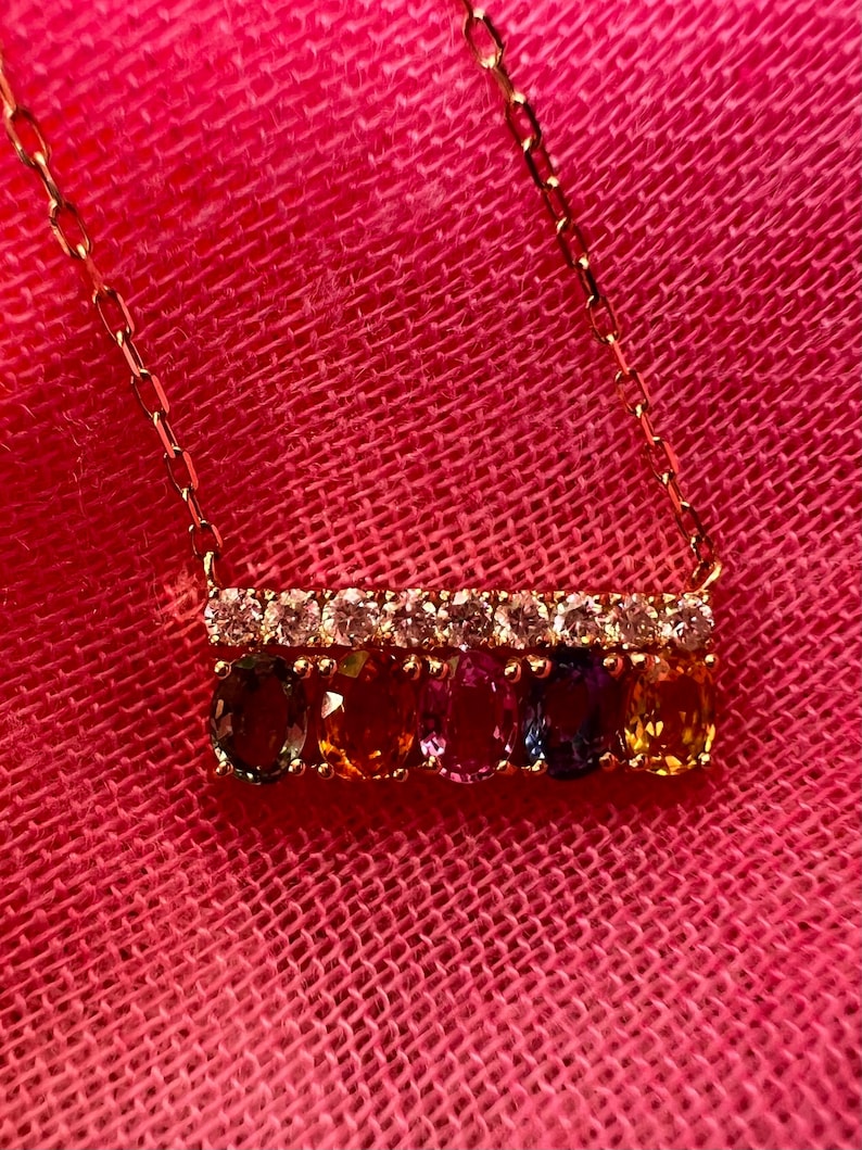 May include: A gold necklace with a bar pendant featuring a row of clear stones above a row of oval-cut gemstones in various colors. The necklace is displayed against a pink background.