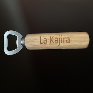 May include: A bottle opener with a light wood handle and a silver metal opener. The text "La Kajira" is engraved on the handle. The opener is designed for removing bottle caps.