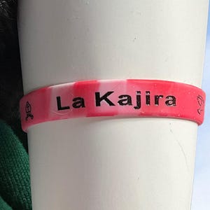 May include: A pink and white striped silicone wristband with the text "La Kajira" in black. The bracelet has a small symbol on each side. The wristband is wrapped around a white cylindrical object.