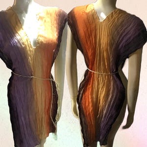 May include: A sleeveless tunic dress featuring a crinkled texture and a vertical gradient color pattern transitioning from deep purple to orange and yellow. The garment has a V-neckline and is cinched at the waist with a thin, light-colored cord.