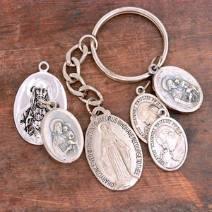 May include: A silver-toned metal keychain featuring a circular ring and a short chain attached to multiple oval-shaped religious pendants. The charms depict various figures, including the Virgin Mary with the text O MARY CONCEIVED WITHOUT SIN PRAY FOR US WHO HAVE RECOURSE TO THEE, and Pope John Paul II with the text BEATUS IOANNES PAULUS PP II.