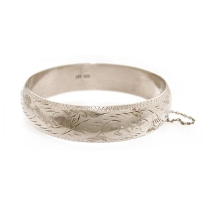 May include: A silver bangle bracelet with an engraved floral design. The bracelet has a clasp with a safety chain. The metal has a shiny finish. The bracelet is a piece of jewelry.