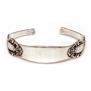 May include: A silver cuff bracelet made from repurposed silverware. The bracelet features a floral design on each end and a smooth, rectangular center. The open design allows for easy wear.