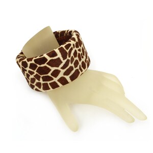 May include: A wide, fabric bracelet with a giraffe print in brown and cream colors. The bracelet is displayed on a mannequin hand against a white background. The cuff-style bracelet is a bold accessory.