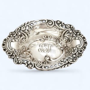 May include: An oval, ornate silver dish with a detailed, scrolling design around the rim. The center features an engraved monogram. The dish has a bright, reflective surface and is likely made of sterling silver.