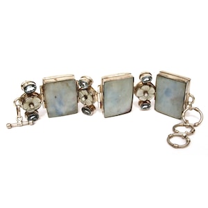 May include: A silver bracelet featuring three rectangular moonstone cabochons alternating with floral-carved mother-of-pearl accents. The design includes small oval blue gemstones and a toggle clasp closure with multiple circular link rings.