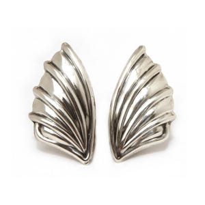 May include: A pair of silver-toned, wing-shaped earrings. Each earring features a fan-like design with multiple curved ridges, creating a textured, reflective surface. The earrings are displayed against a white background.