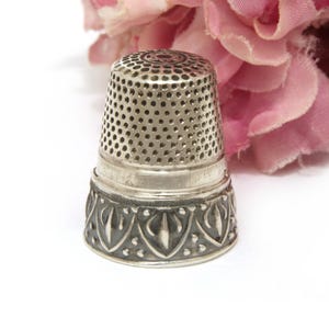 May include: A silver thimble with a perforated top and decorative embossed base. The thimble is set against a soft pink floral background. This sewing tool is designed to protect the finger while hand sewing.