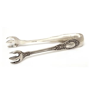 May include: Silver sugar tongs with ornate detailing. The tongs feature a decorative handle with an engraved design and a monogram. The ends have three-pronged forks for gripping sugar cubes. The tongs are made of sterling silver.