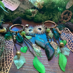 Puede incluir: Pendientes de aro dorados con colgantes de hojas y cristales. Los colgantes incluyen hojas verdes, transparentes y doradas, cristales verdes y pequeñas cuentas doradas. El fondo incluye musgo, flores secas y una nota con texto.