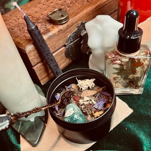 Può includere: Un assortimento di oggetti spirituali, tra cui una candela nera, un piccolo contenitore nero pieno di erbe e cristalli, una bottiglia di vetro con un contagocce e una candela bianca a forma di torso. Gli oggetti sono disposti su una superficie di velluto verde.