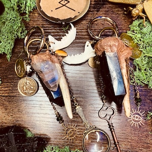 May include: Two keychains with bone, crystal, and metal accents, alongside a vintage magnifying glass and key. A small, round, antique-style compact with a symbol is also present. The keychains feature gold-colored rings and chains, with a sun-shaped charm.